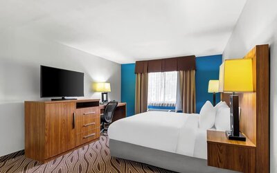 Modern hotel guest room with a king bed, white bedding, wooden furniture, flat-screen television, work desk and chair, bedside lamp, window, and blue accent wall.