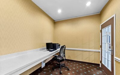Hotel business center with a long white work counter, desktop computer, printer, ergonomic office chair, recessed ceiling lights, and a glass-paneled door.