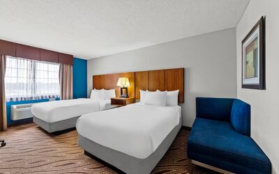 Bright hotel guest room with two beds dressed in white linens, wooden headboards, bedside lamp, large window, blue accent wall, patterned carpet, and blue upholstered chaise.