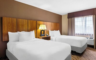Spacious hotel room with two queen beds, crisp white bedding, warm wooden headboards, central bedside table and lamp, large curtained window, and patterned carpeting.