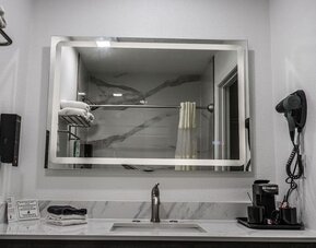 Bathroom vanity with a large illuminated mirror, marble-look backsplash, sink, hairdryer, towels, and in-room coffee setup at Western Inn.