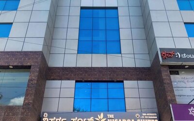 Multistory exterior of The Nisarga Suites in Bangalore featuring reflective blue glass windows, silver facade panels, brown stone accents, and hotel signage.