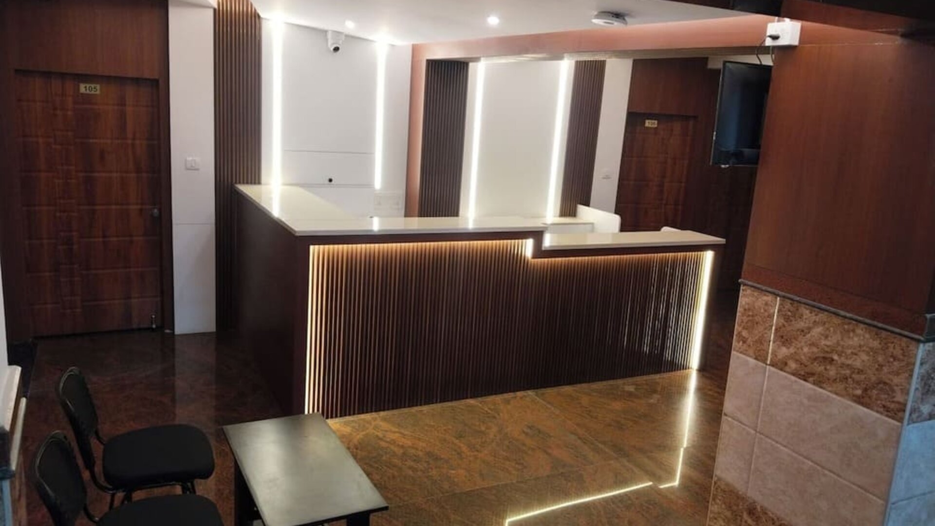 Modern hotel reception area featuring a dark wooden front desk with white countertops, decorative LED lighting, polished flooring, and guest seating.
