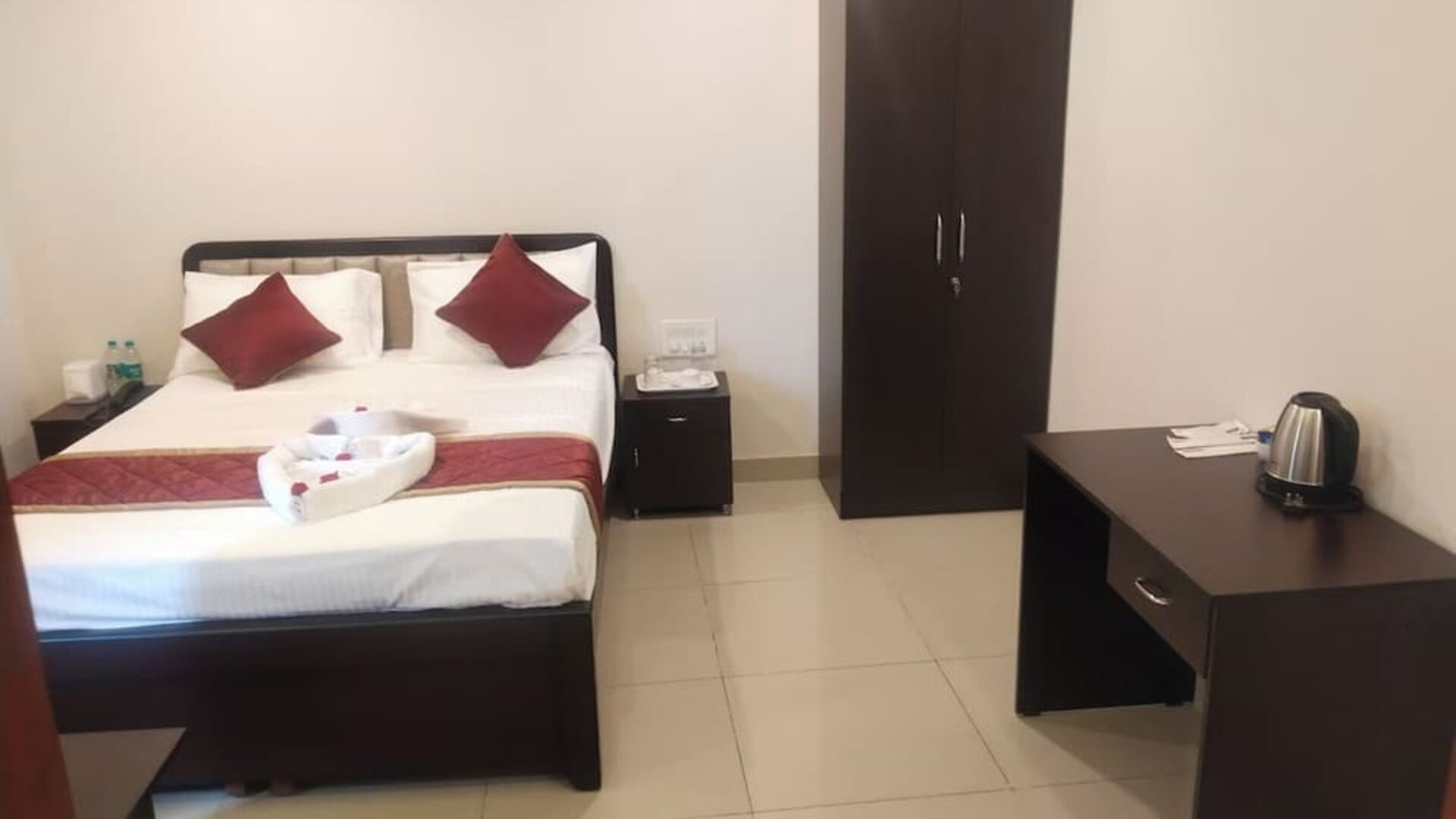 Bright double bed guest room featuring white bedding, burgundy accents, a wooden wardrobe, bedside cabinets, and a refreshment station with an electric kettle.