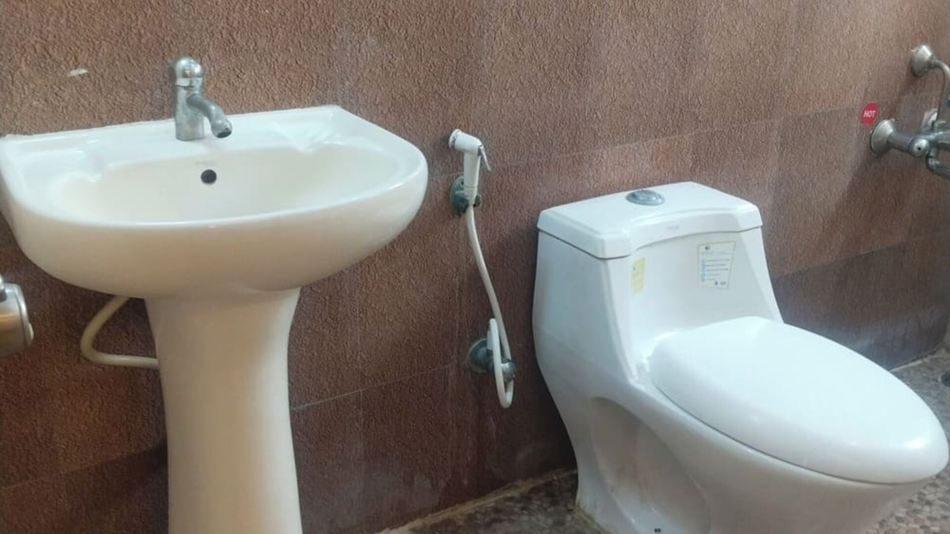 Private hotel bathroom featuring a white pedestal sink, modern toilet, handheld bidet sprayer, wall-mounted shower fixtures, large mirror, and patterned tile flooring.