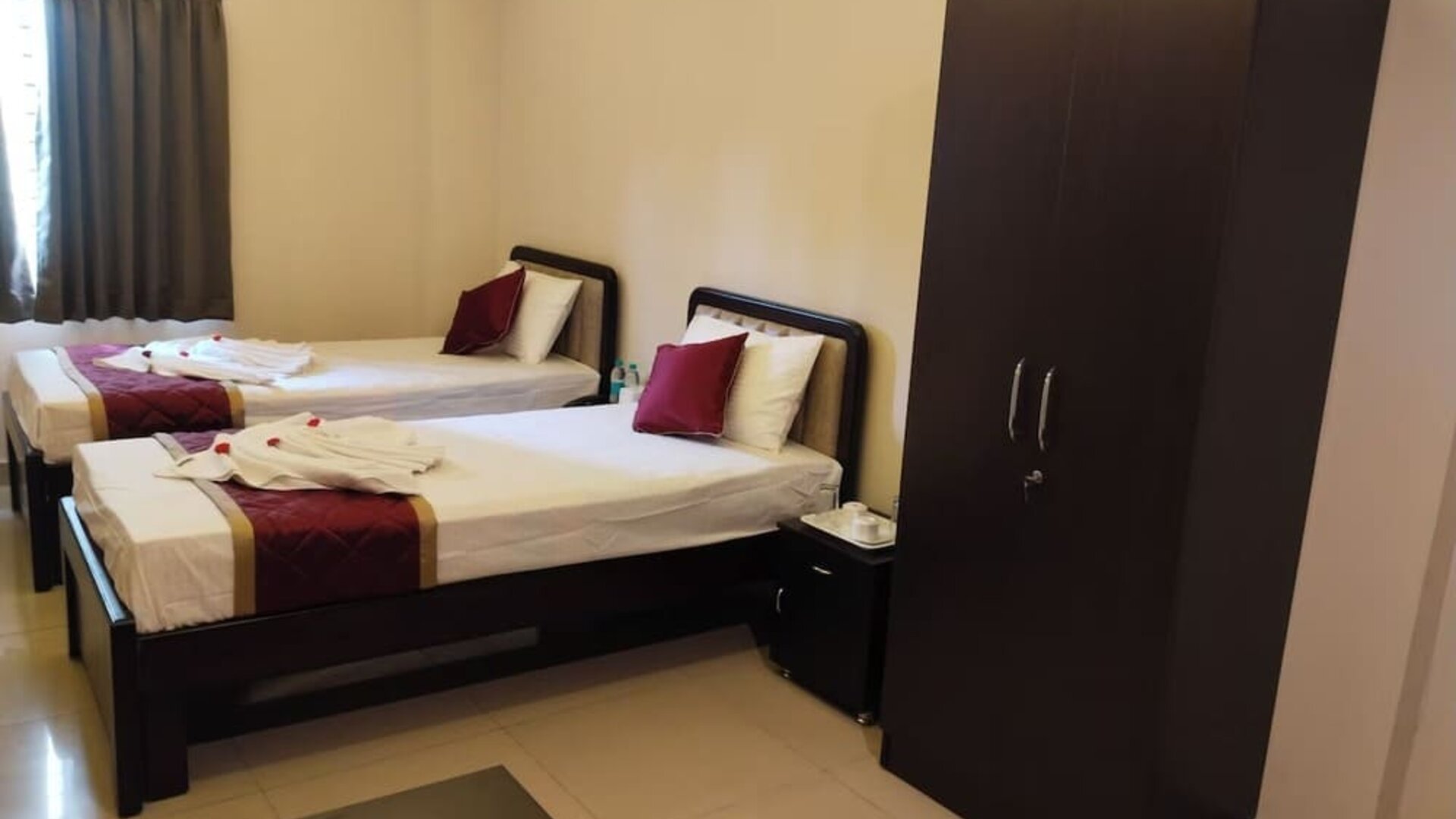 Twin bed hotel room featuring white bedding, burgundy accents, decorative towel arrangements, dark wooden furniture, a spacious wardrobe, and tiled flooring.