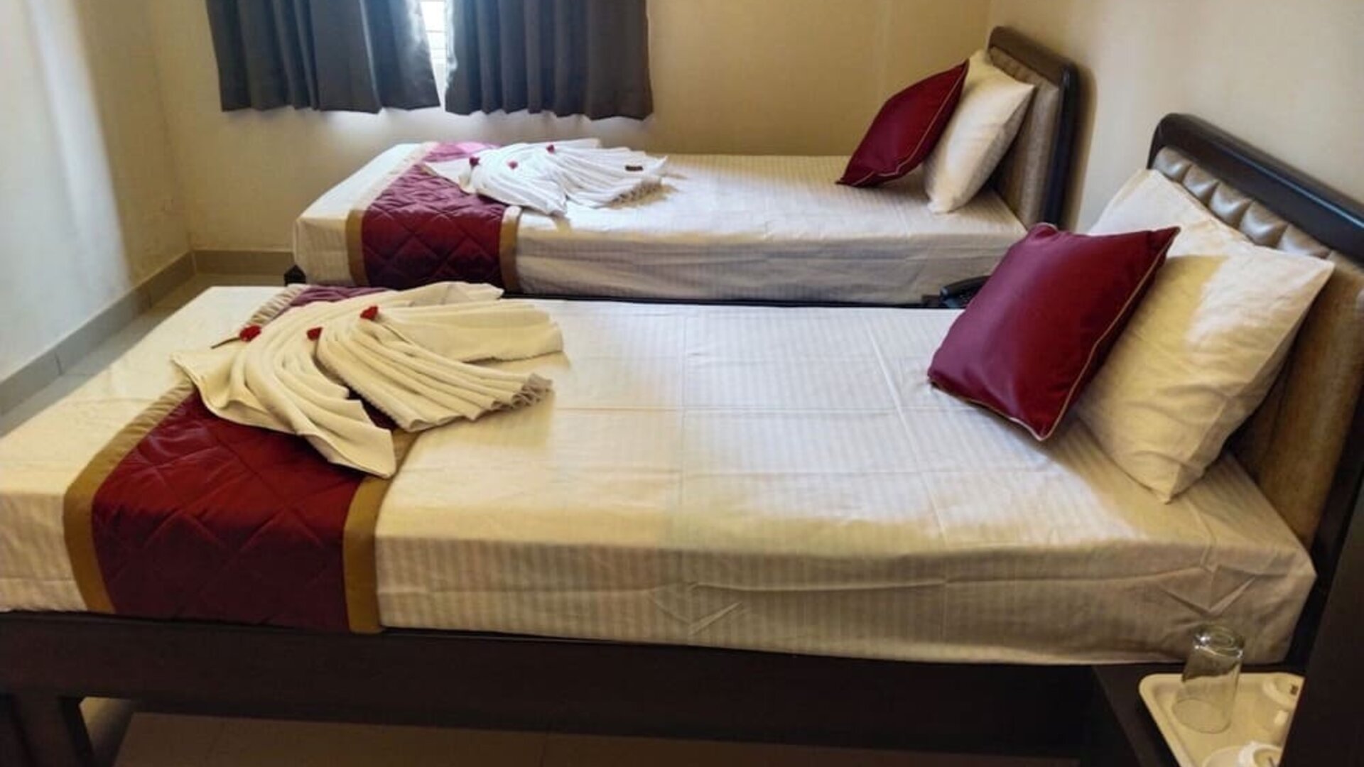 Neatly furnished twin bed guest room featuring white linens, burgundy quilted runners, decorative towel arrangements, dark wooden bed frames, and a curtained window.