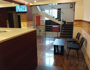 Welcoming hotel lobby featuring a polished reception counter, black guest chairs, coffee table, staircase with stainless steel railings, and glossy tiled flooring.