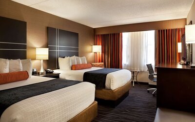 Guest room with two beds, crisp white bedding, dark bed runners, orange accent cushions, striped curtains, bedside lamps, and a work desk at Best Western Plus BWI Airport Arundel Mills.