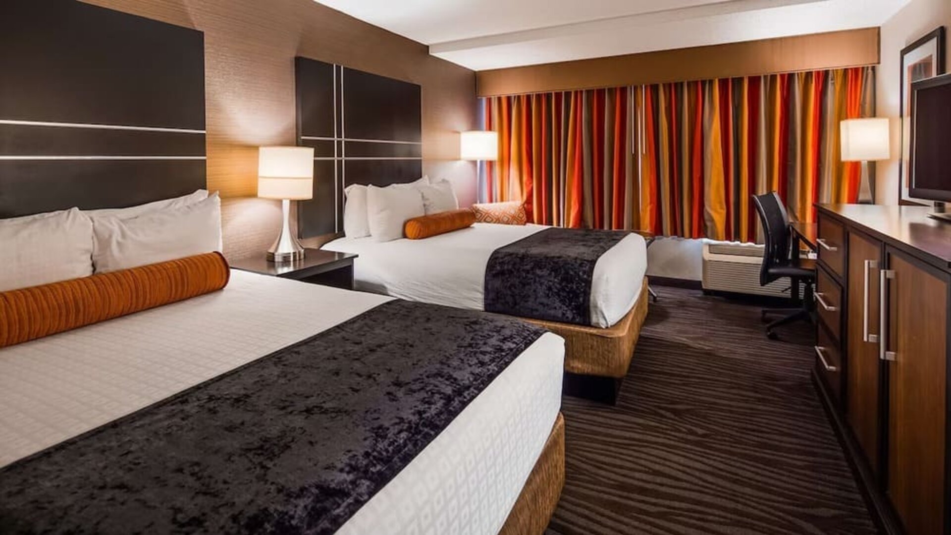 Guest room with two beds, white bedding, dark bed runners, orange accent cushions, striped curtains, bedside lamps, and a work desk at Best Western Plus BWI Airport Arundel Mills.