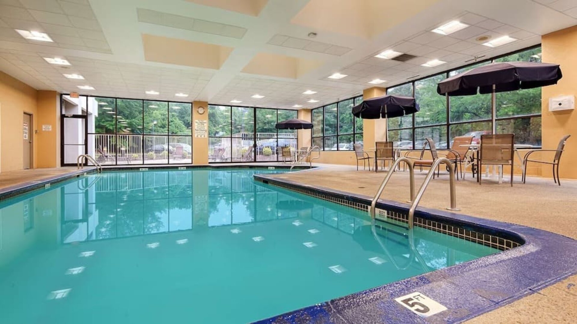 Indoor swimming pool with clear turquoise water, surrounding deck, metal handrails, large windows, and outdoor greenery visible through the glass at Best Western Plus BWI Airport Arundel Mills.