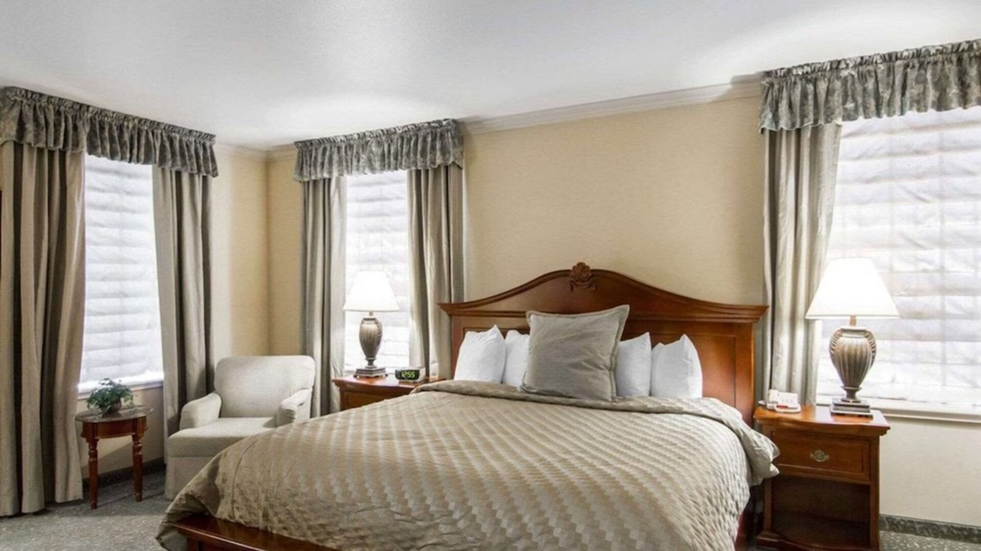 King guest room with natural window light, a traditional wooden bed, beige patterned bedding, bedside lamps, upholstered armchairs, and neutral curtains at Liberty Hotel Cleburne, an Ascend Hotel Collection Member.