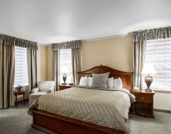 King guest room with natural window light, a traditional wooden bed, beige patterned bedding, bedside lamps, upholstered armchairs, and neutral curtains at Liberty Hotel Cleburne, an Ascend Hotel Collection Member.
