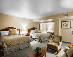 Spacious guest room featuring two beds with beige patterned bedding, wooden headboards, matching bedside tables, upholstered seating, and a carpeted floor at Liberty Hotel Cleburne, an Ascend Hotel Collection Member.