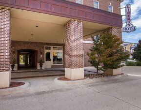 Exterior entrance of Liberty Hotel Cleburne, featuring a red-brick facade, covered porte-cochère, brick columns, landscaped planters, and the hotel's vertical sign.
