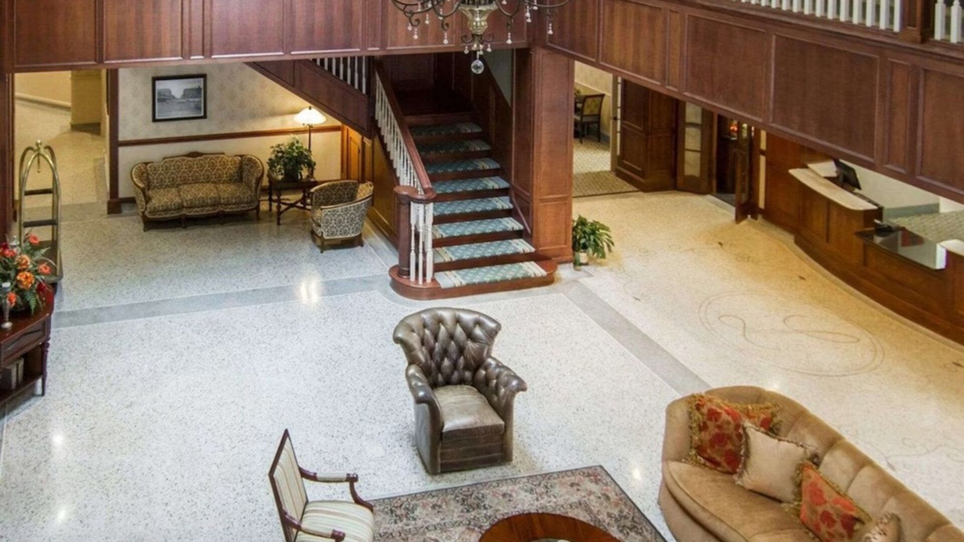 Hotel lobby lounge featuring leather armchairs, upholstered sofas, wooden furniture, a decorative chandelier, a central staircase, and a polished terrazzo floor at Liberty Hotel Cleburne, an Ascend Hotel Collection Member.