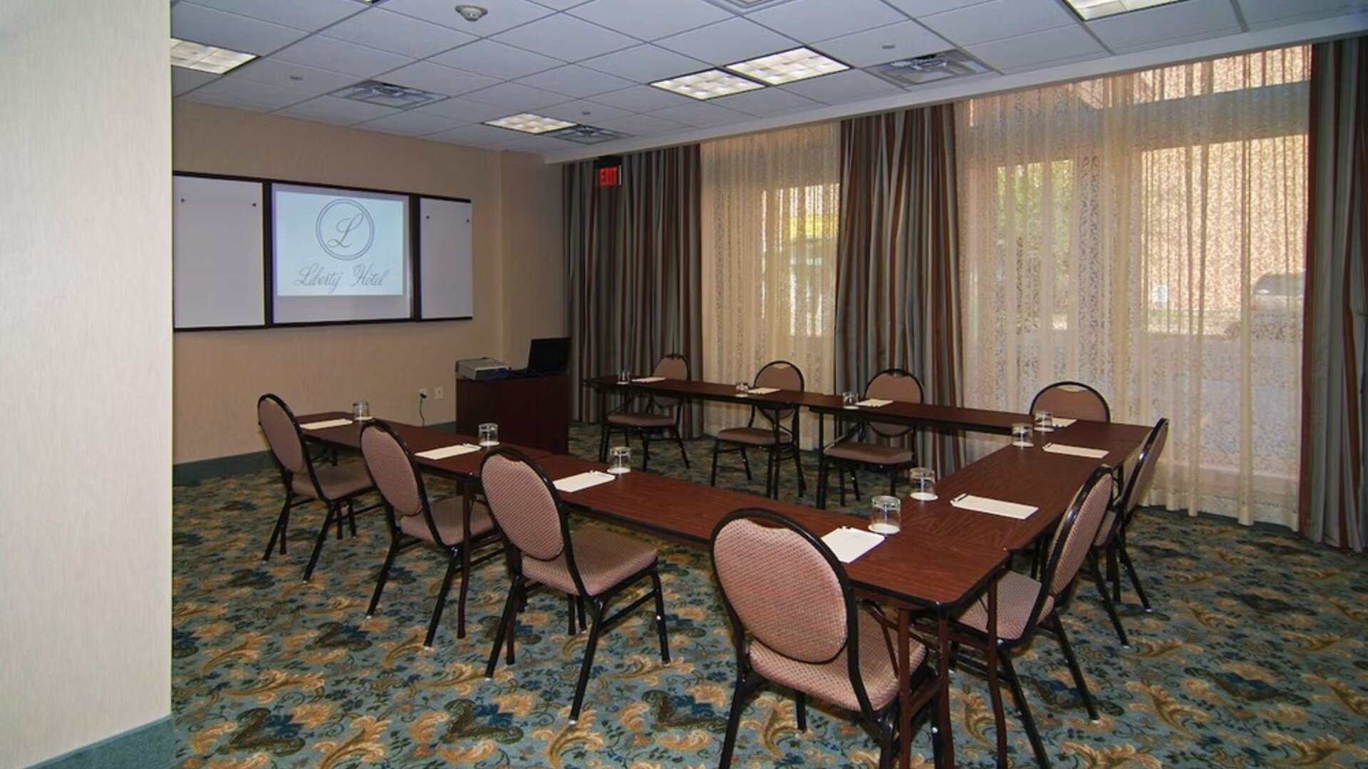 Meeting room arranged with conference tables and chairs in a U-shaped layout, a projection screen, presentation equipment, large curtained windows, and patterned carpeting at Liberty Hotel Cleburne, an Ascend Hotel Collection Member.