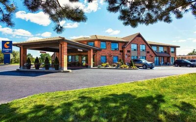 Exterior view of Comfort Inn Alma, showcasing its red-brick facade, covered entrance, blue hotel signage, parking area, and landscaped lawn.
