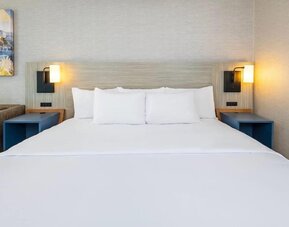 Close-up of a king bed featuring crisp white bedding, multiple pillows, a light wood headboard, modern wall sconces, and blue bedside shelving at Comfort Inn Alma.