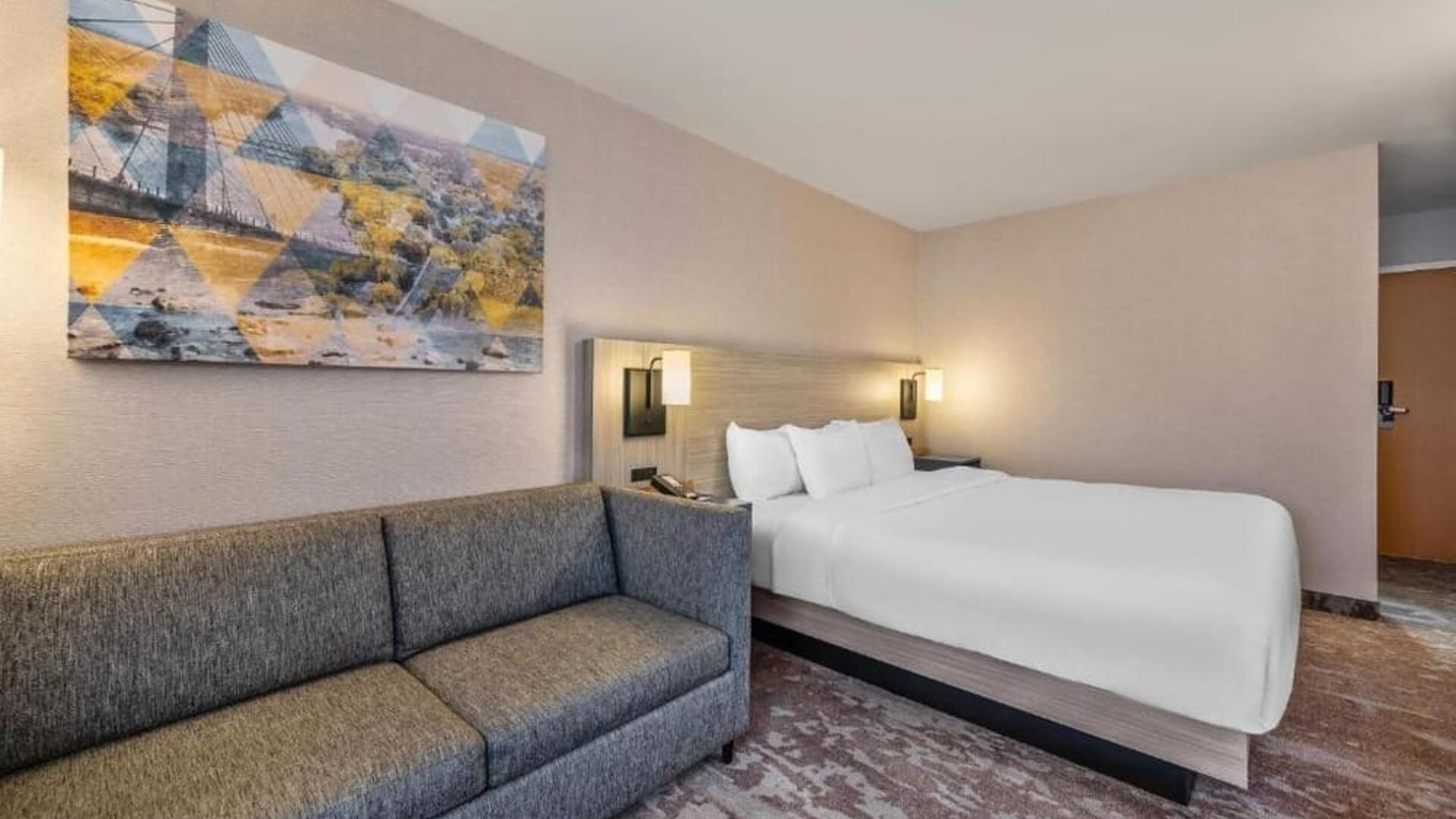 Spacious king guest room with a large bed, crisp white bedding, a gray sofa, abstract wall art, and patterned carpeting at Comfort Inn Alma.
