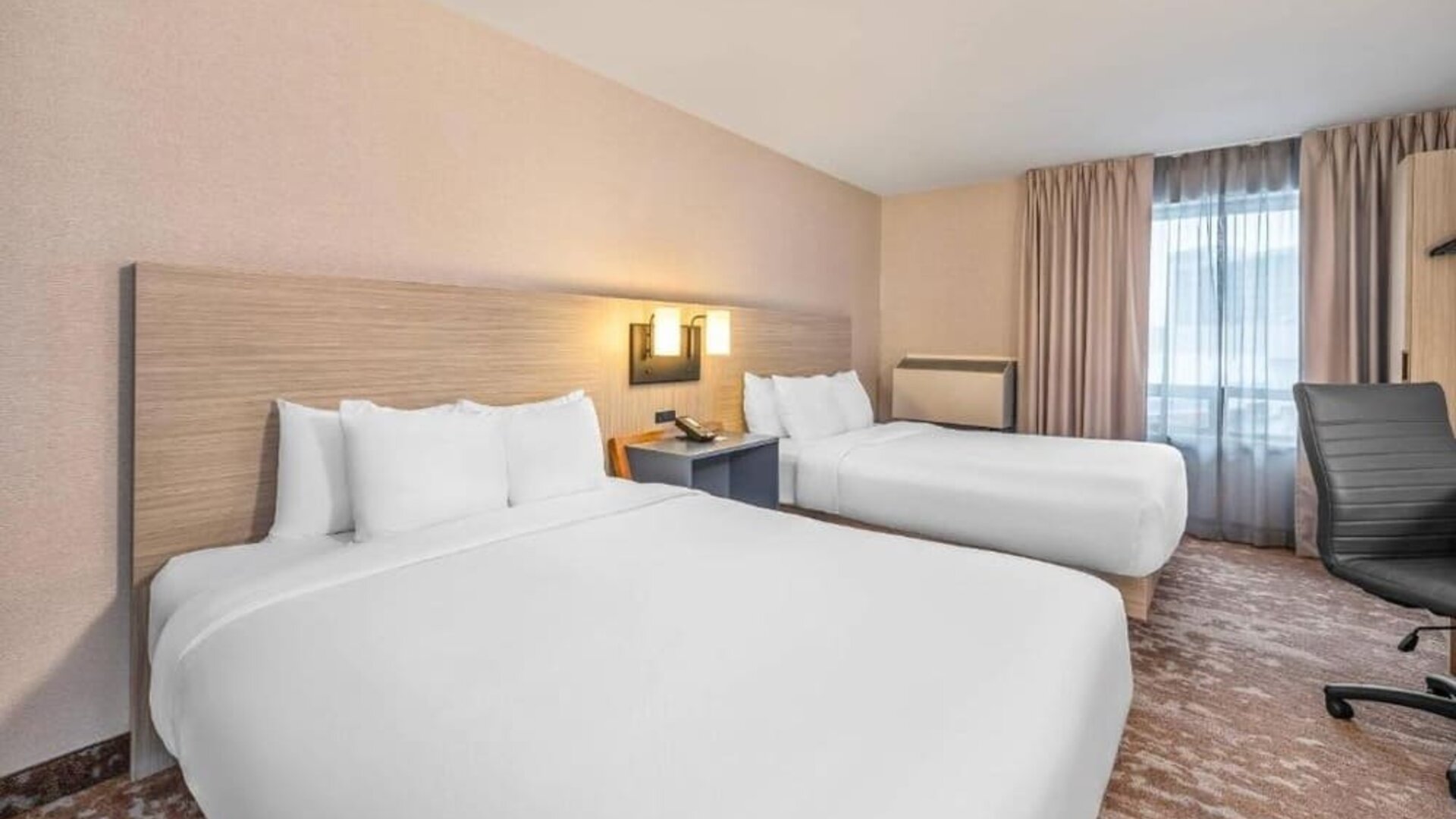 Guest room with two beds featuring white bedding, light wood furnishings, modern wall sconces, a desk and office chair, and neutral decor at Comfort Inn Alma.
