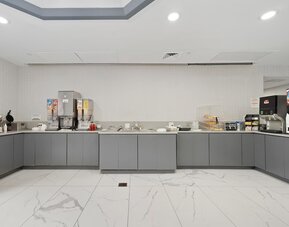 Breakfast buffet area with gray cabinetry, cereal dispensers, beverage machines, serving counters, and a white marble-patterned tile floor at Chicago Club Inn & Suites.