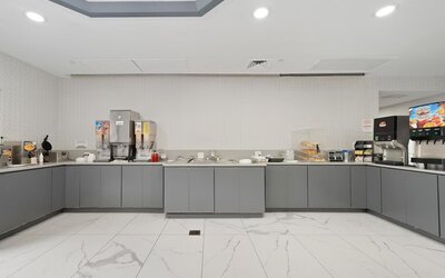 Breakfast buffet area with gray cabinetry, cereal dispensers, beverage machines, serving counters, and a white marble-patterned tile floor at Chicago Club Inn & Suites.