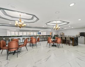 Hotel dining and breakfast area with marble-patterned flooring, orange upholstered chairs, tables, a breakfast counter, and beverage stations at Chicago Club Inn & Suites.