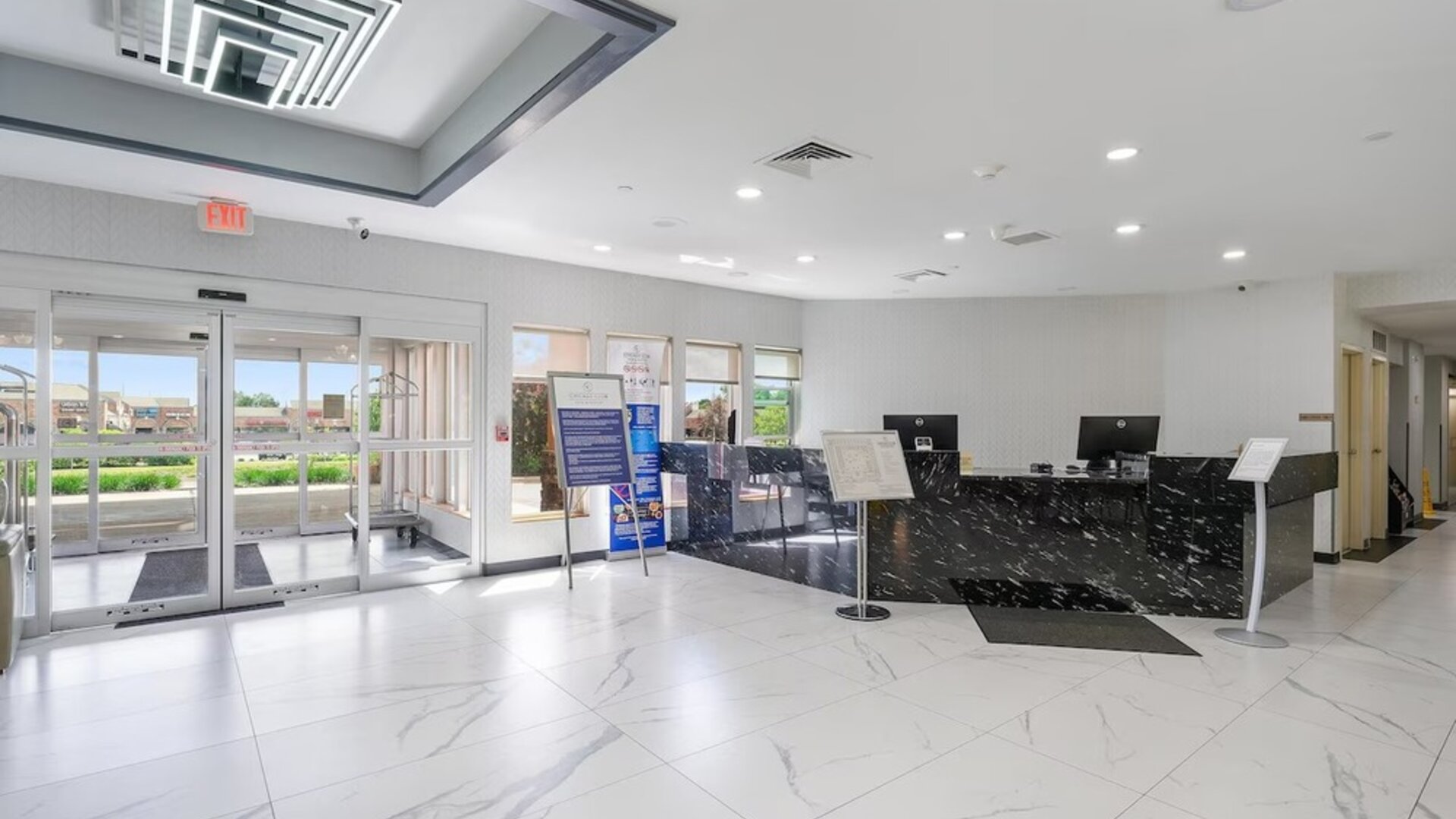 Hotel reception area featuring a black marble-patterned front desk, computer workstations, glass entrance doors, and a bright tiled lobby at Chicago Club Inn & Suites.