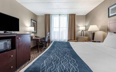 A king guest room features white bedding with a blue patterned runner, a flat-screen television, microwave, desk and office chair at Comfort Inn & Suites Glen Mills – Concordville.