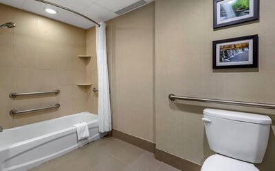 A beige-tiled bathroom with a bathtub, white shower curtain, toilet and metal grab bars along the walls at Comfort Inn & Suites Glen Mills – Concordville.