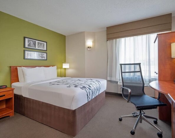 Bright Sleep Inn king room featuring a large bed with white bedding and gray floral detailing, green accent wall, framed artwork, wooden bedside tables, modern lamps, spacious work desk, black mesh office chair, and a window with sheer curtains.