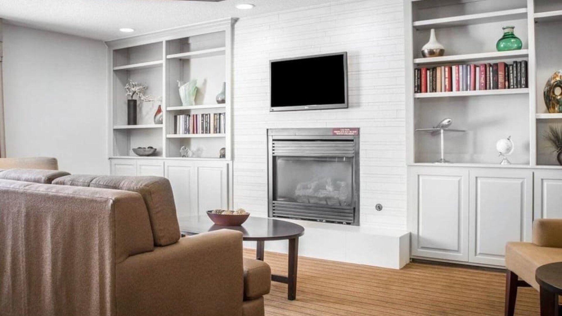 Comfortable Sleep Inn lobby lounge featuring beige upholstered sofas and armchairs, a central fireplace with a wall-mounted flat-screen television, built-in white bookshelves displaying books and decorative accessories, and a round coffee table.