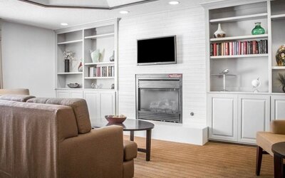 Comfortable Sleep Inn lobby lounge featuring beige upholstered sofas and armchairs, a central fireplace with a wall-mounted flat-screen television, built-in white bookshelves displaying books and decorative accessories, and a round coffee table.