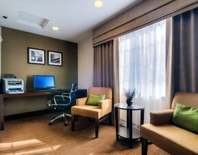 Cozy hotel business center featuring a desktop computer, printer, dark wooden workstation, ergonomic office chair, two beige upholstered armchairs with green accent pillows, a round side table, and a large window providing natural light.