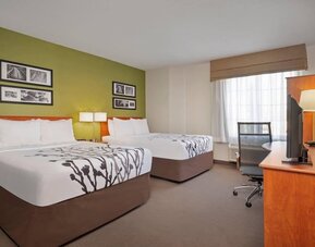 Spacious Sleep Inn guest room featuring two beds with crisp white linens and decorative gray floral patterns, a green accent wall with framed black-and-white artwork, wooden furniture, a flat-screen television, work desk, ergonomic office chair, and a lar