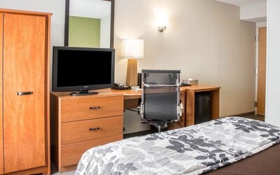 Sleep Inn guest room featuring a wooden dresser with flat-screen television, spacious work desk with black ergonomic office chair, large wall mirror, bedside lamp, wooden wardrobe, mini refrigerator, and a bed with brown bedding and a gray floral-patterne