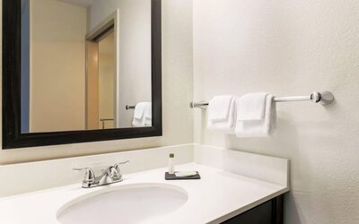 Clean hotel bathroom vanity featuring a white countertop with an integrated sink, chrome faucet, large black-framed mirror, neatly folded white hand towels, wall-mounted towel rack, and complimentary toiletries.