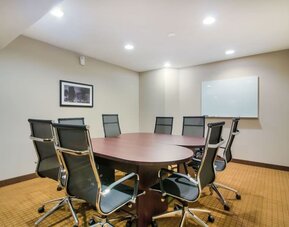 Professional hotel meeting room featuring a large oval wooden conference table surrounded by black ergonomic office chairs, a wall-mounted whiteboard, framed artwork, beige carpeting, and recessed ceiling lighting.
