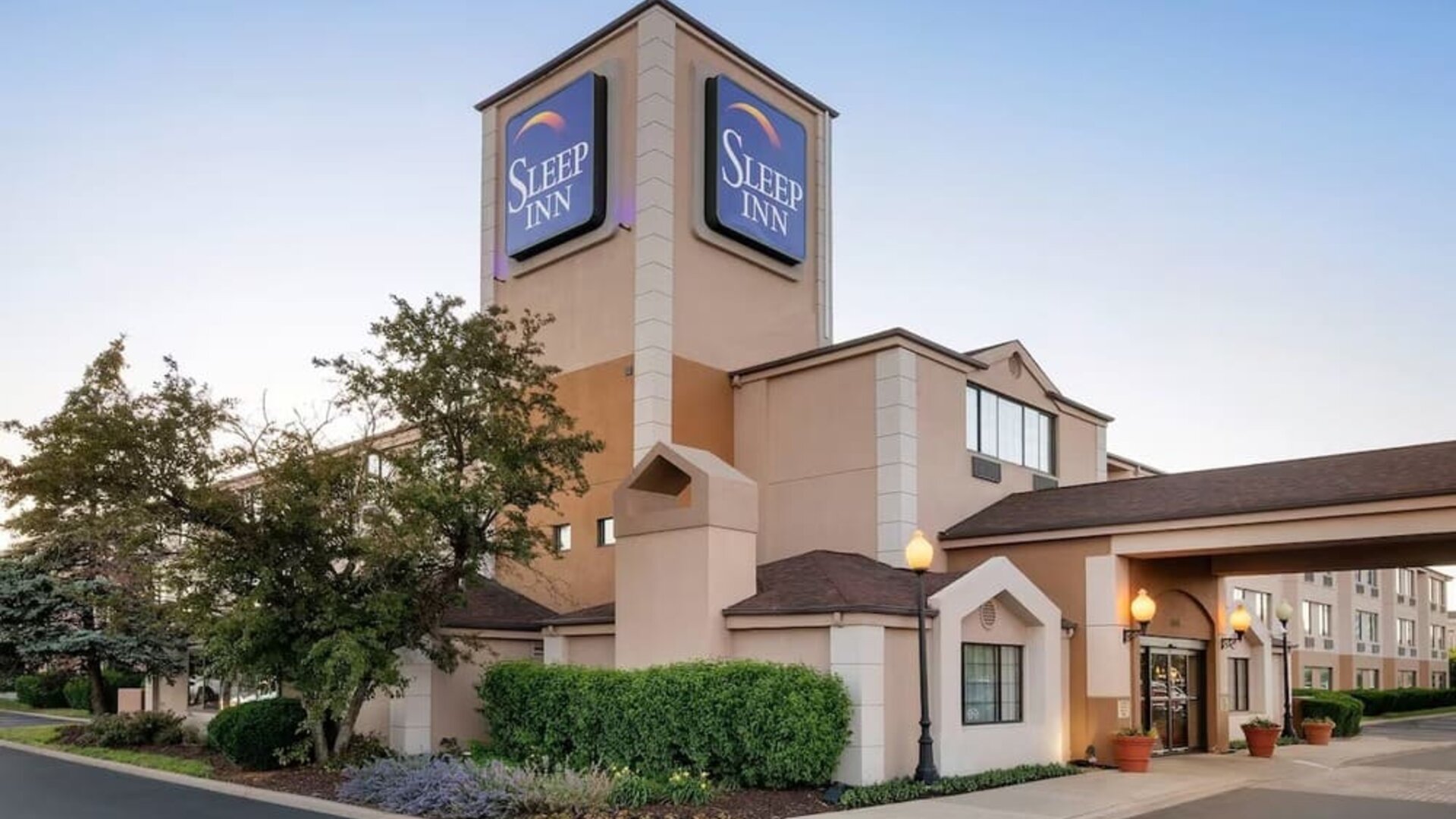 Exterior view of the Sleep Inn hotel at dusk featuring a multi-story beige building, illuminated blue Sleep Inn signage, covered main entrance, landscaped trees and shrubs, decorative lampposts, and a paved driveway.