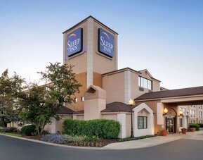 Exterior view of the Sleep Inn hotel at dusk featuring a multi-story beige building, illuminated blue Sleep Inn signage, covered main entrance, landscaped trees and shrubs, decorative lampposts, and a paved driveway.