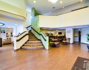 Spacious Sleep Inn lobby featuring a central carpeted staircase with green floral-patterned accent walls, wood-look tile flooring, comfortable lounge seating, an adjacent dining area with wooden tables and chairs, and bright overhead lighting.