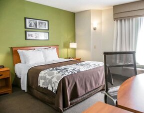 omfortable Sleep Inn king room featuring a large bed with white pillows, brown bedding, and a gray floral-patterned runner, green accent wall with framed artwork, wooden bedside tables, reading lamps, and a work desk beside a curtained window.
