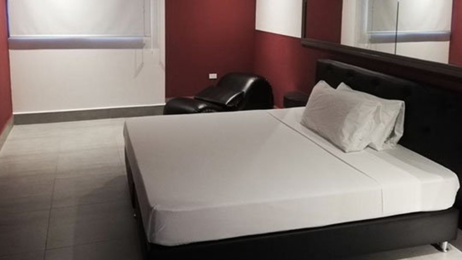 A guest room with a double bed, white linens, a black upholstered headboard, burgundy walls, tiled flooring and a black lounge chair at Residencial & Hotel Palazzo.