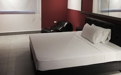 A guest room with a double bed, white linens, a black upholstered headboard, burgundy walls, tiled flooring and a black lounge chair at Residencial & Hotel Palazzo.
