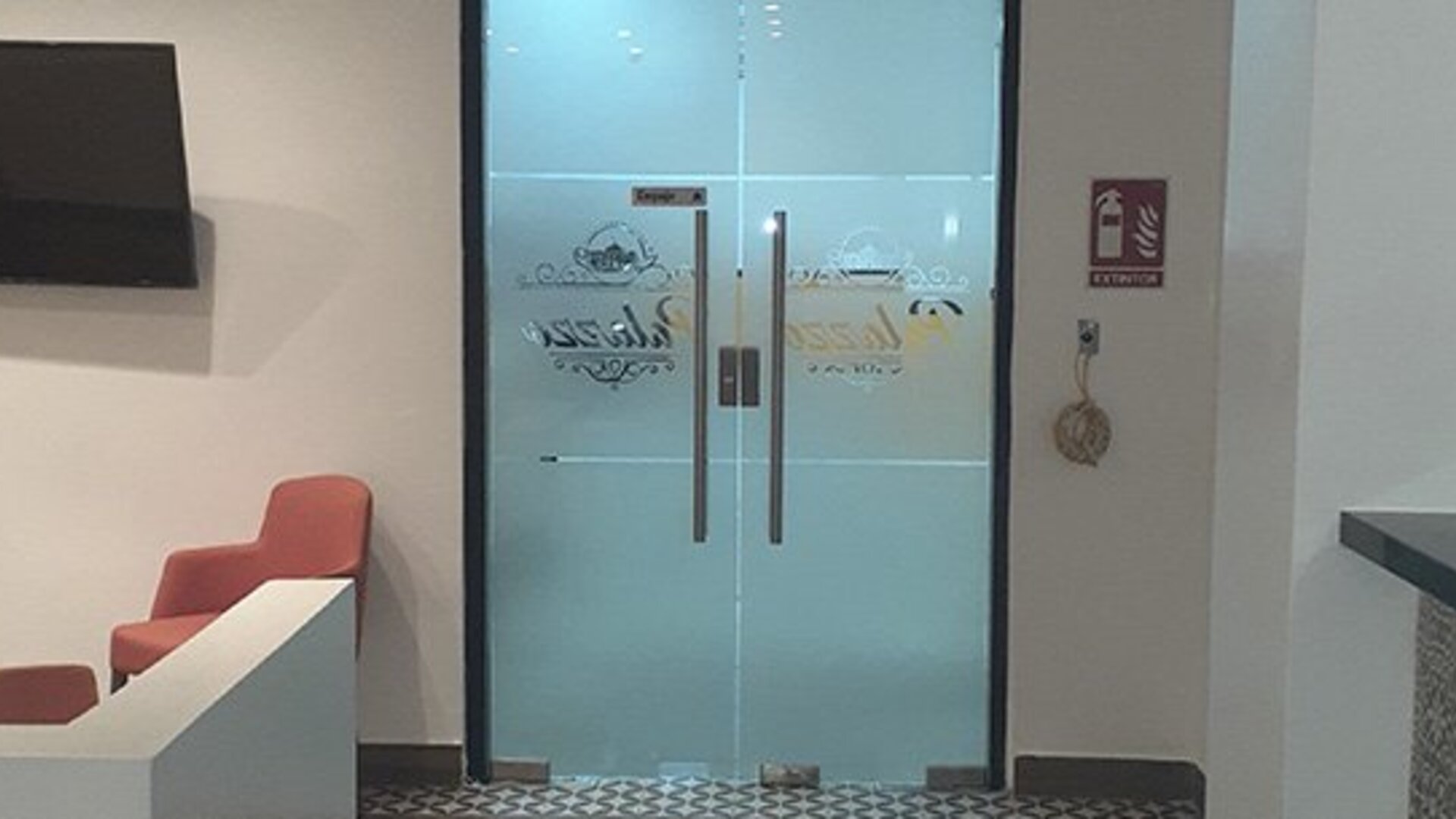 A reception area with frosted glass double doors, a wall-mounted television, coral chairs and a dark countertop at Residencial & Hotel Palazzo.