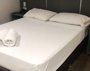 A double bed with white bedding and rolled towels, a dark bedside table with a remote control and a burgundy accent wall at Residencial & Hotel Palazzo.
