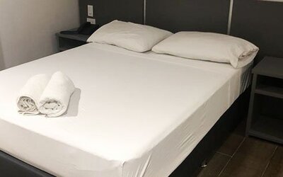A double bed with white bedding and rolled towels, a dark bedside table with a remote control and a burgundy accent wall at Residencial & Hotel Palazzo.