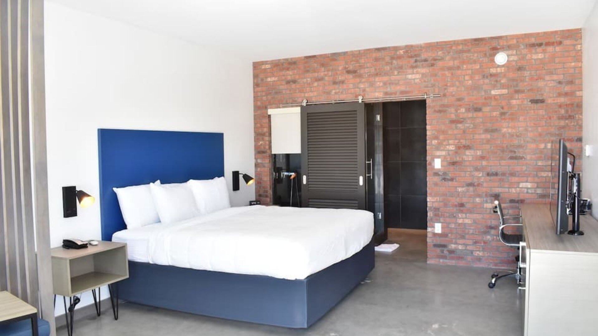 A spacious guest room with a king bed, white linens, a blue upholstered headboard, exposed brick accent wall, desk and office chair at Hotel Bella.