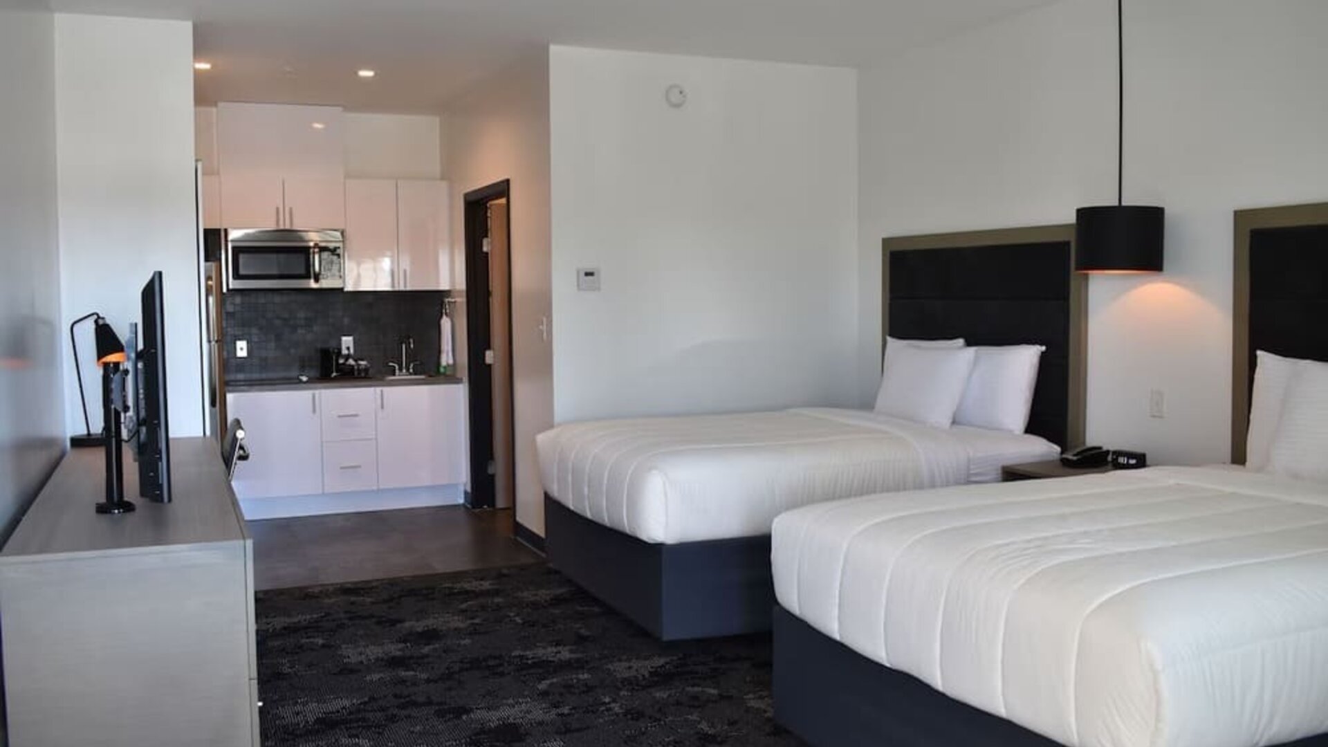 A guest room with two beds dressed in white bedding, black headboards, bedside tables, pendant lighting and a kitchenette in the background at Hotel Bella.